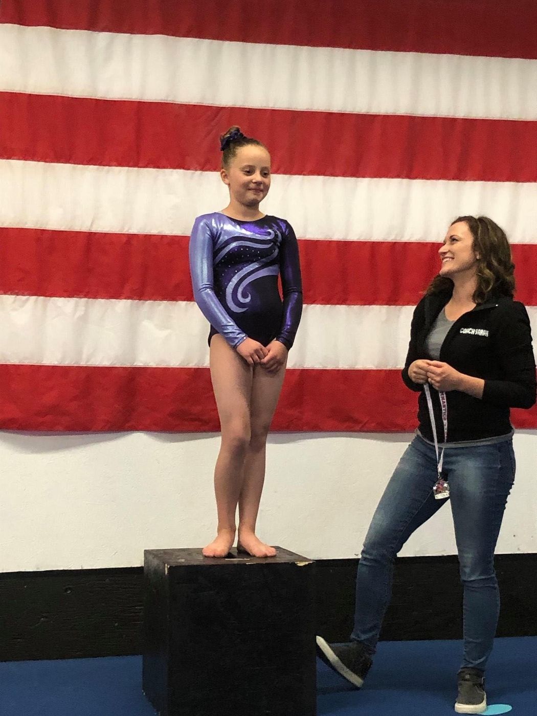 A young gymnast in a purple leotard standing on an awards podium with a coach beside her in front of an American flag.
