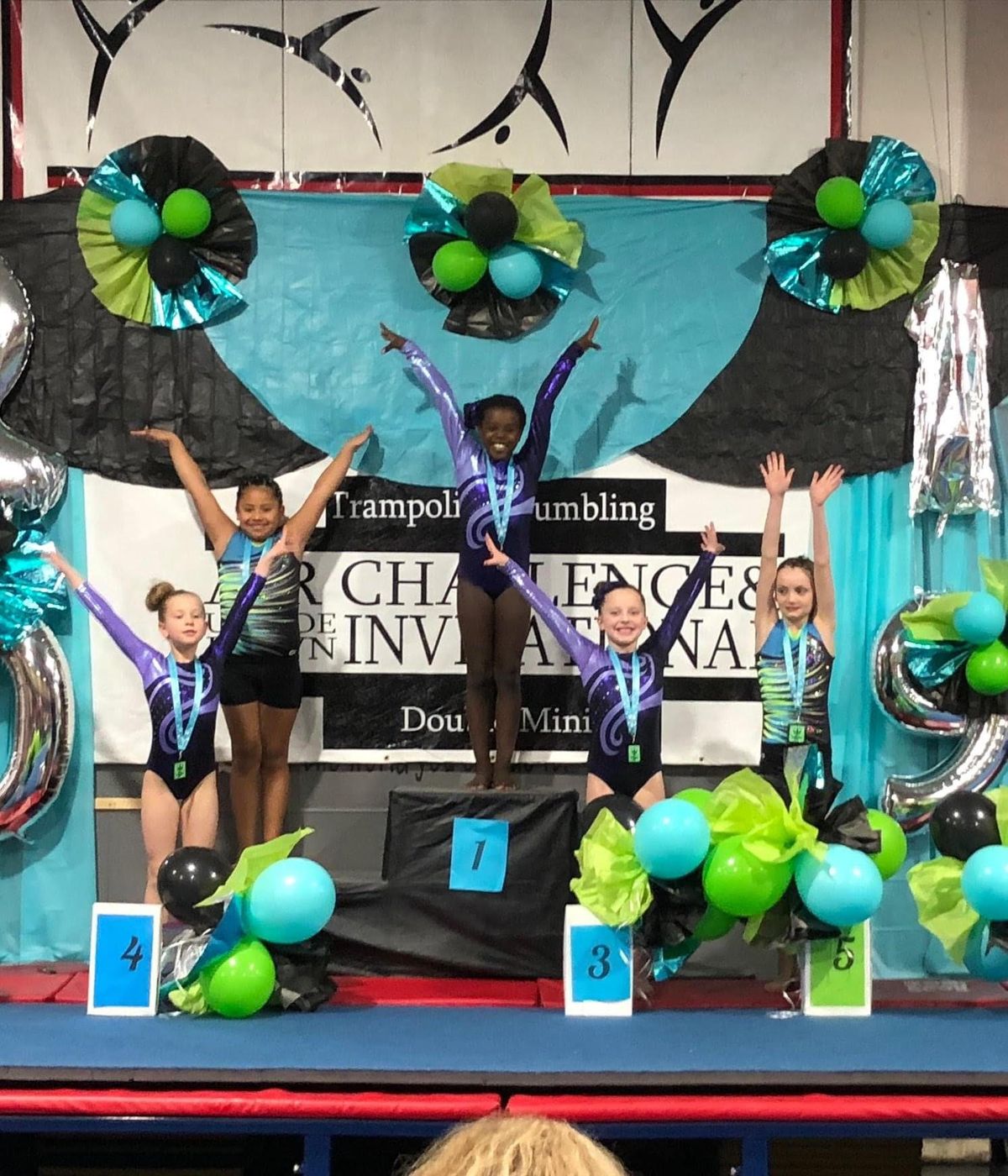 Young gymnasts posing with arms raised on a decorated competition podium.