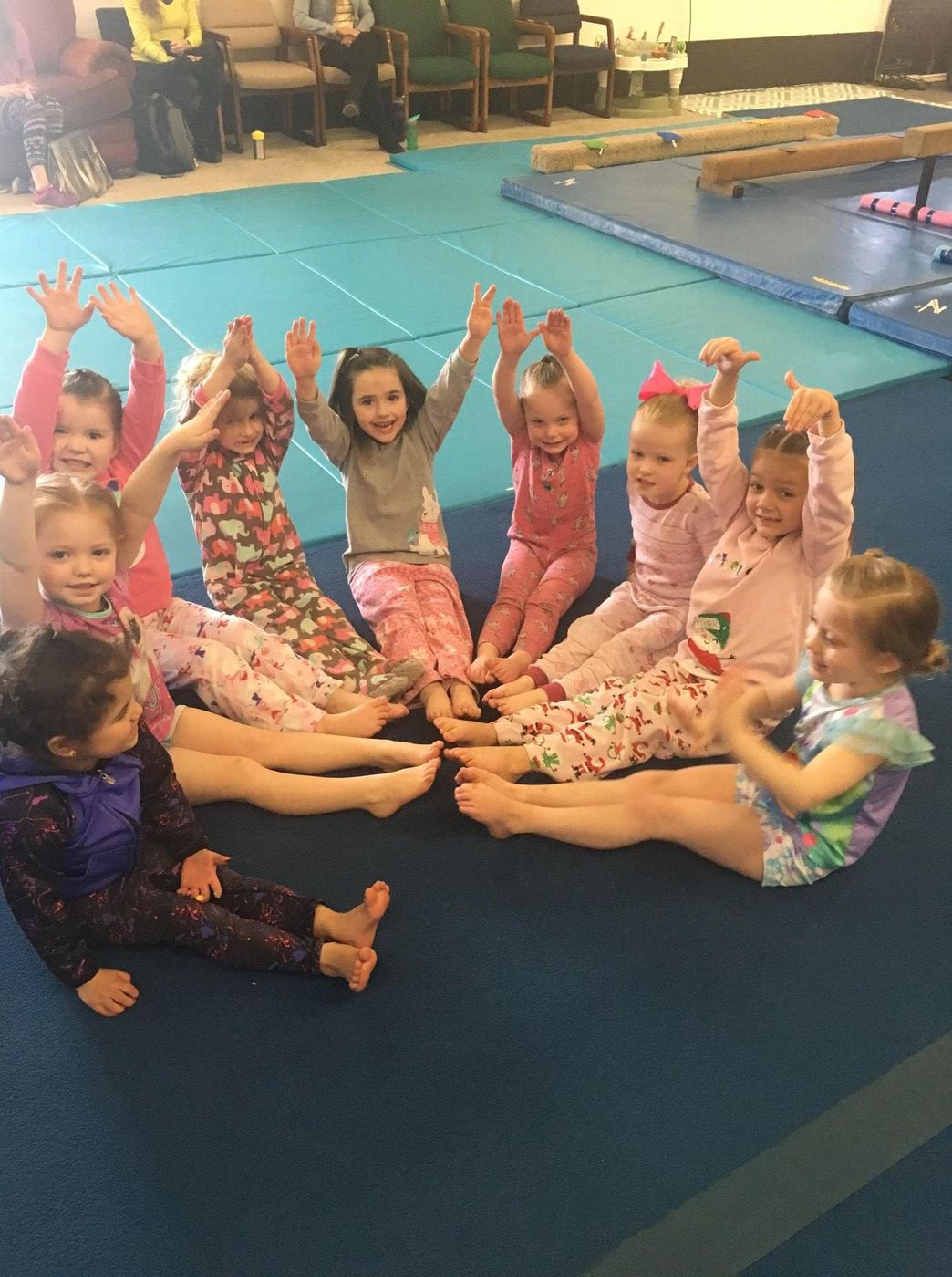 A row of young children sitting on the gym floor with hands raised during a class.