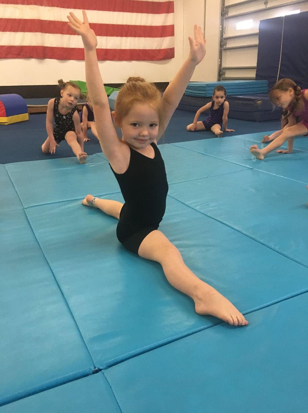A young gymnast doing the splits with arms raised during a floor class.