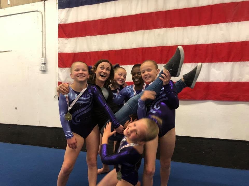 A coach and young gymnasts in purple leotards laughing while lifting a teammate in front of an American flag.