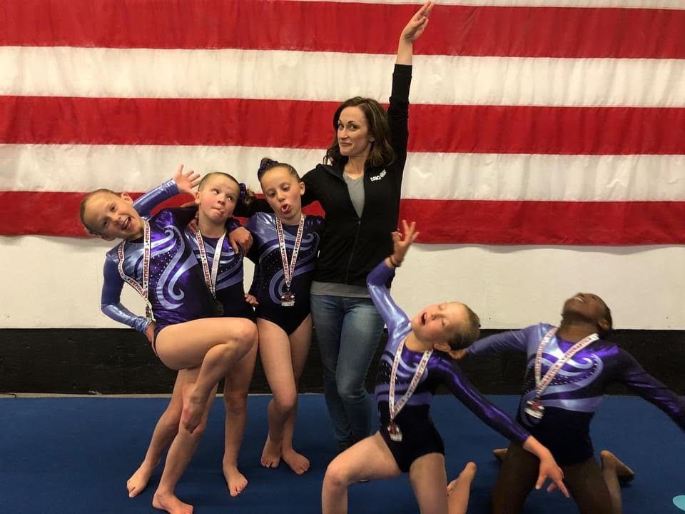 A coach and young gymnasts in purple leotards strike playful poses in front of an American flag.
