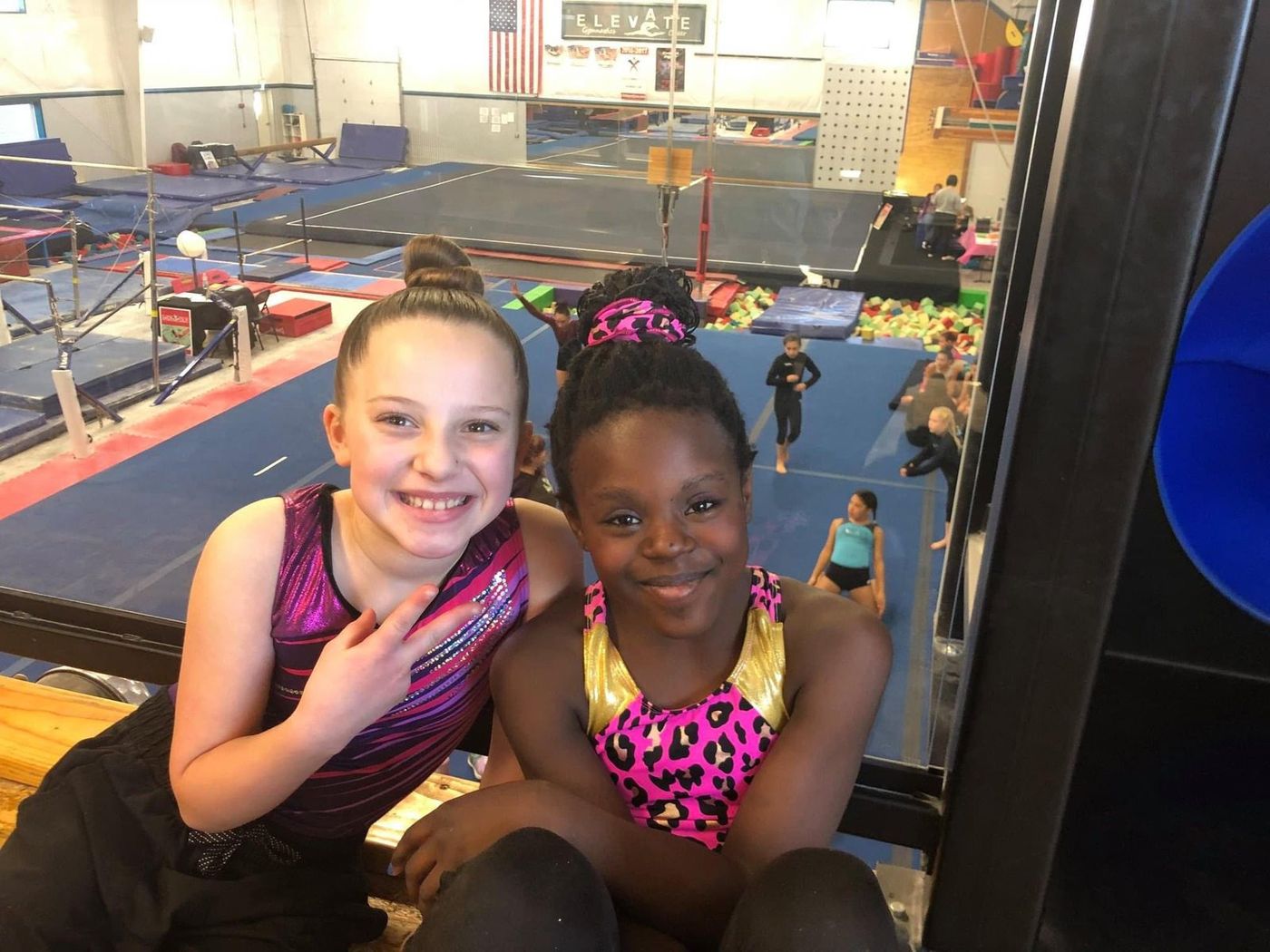 Two smiling young gymnasts posing together above a busy gym training floor.