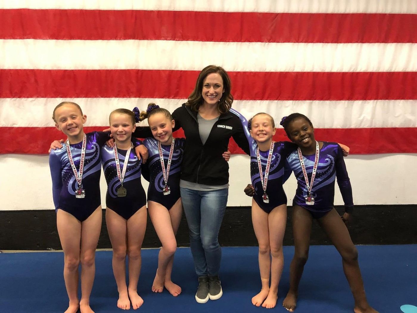 A coach posing with young gymnasts wearing medals in front of an American flag.