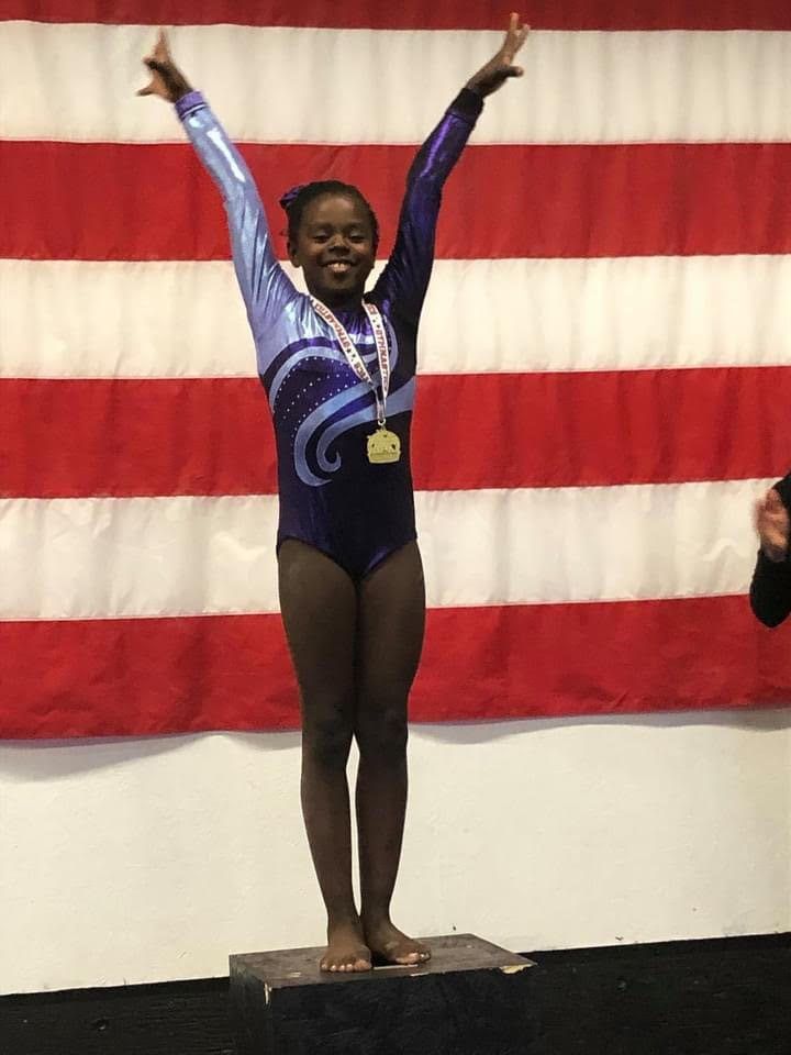 A young gymnast with a medal raising her arms on an awards podium in front of an American flag.