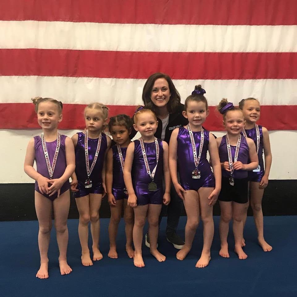 A coach posing with seven young gymnasts in purple leotards and medals in front of an American flag.
