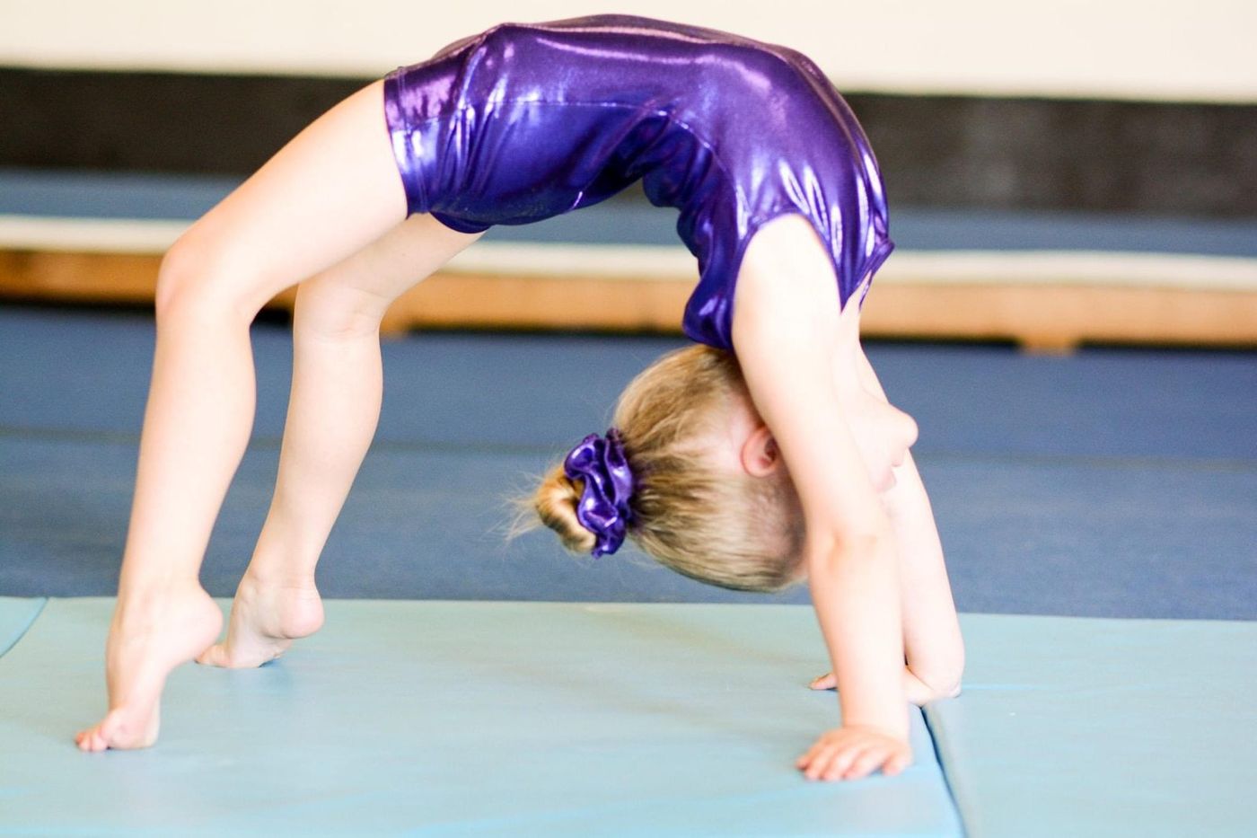 A young girl in a purple leotard doing a backbend bridge on a gym mat.