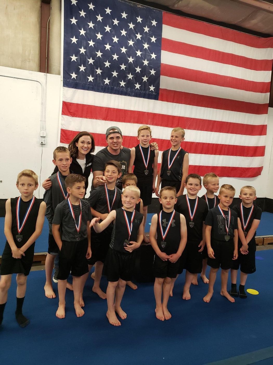 Two coaches posing with a large group of boys wearing medals in front of an American flag.