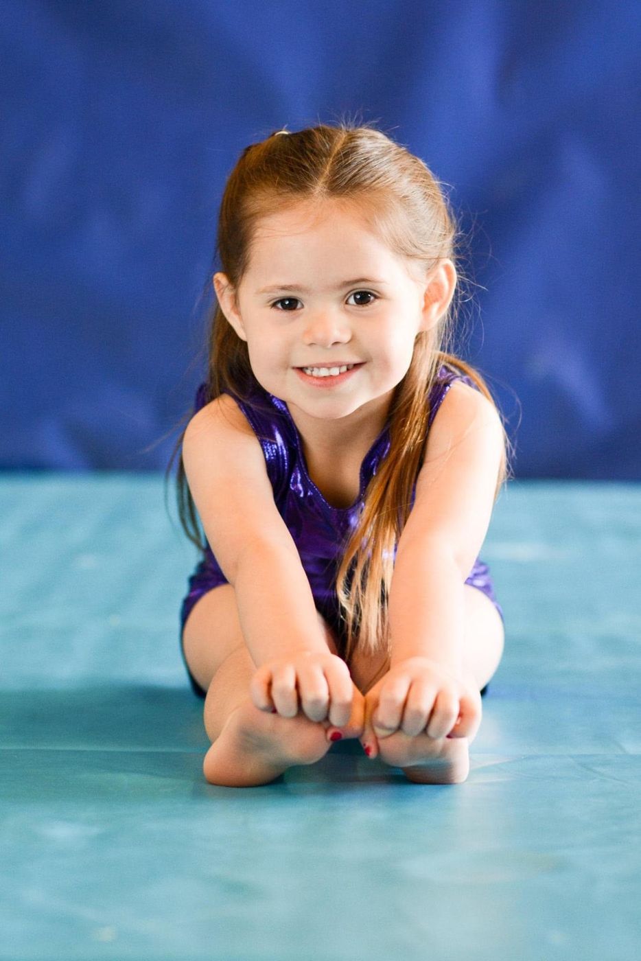 A young girl in a purple leotard sitting and stretching toward her toes against a blue backdrop.
