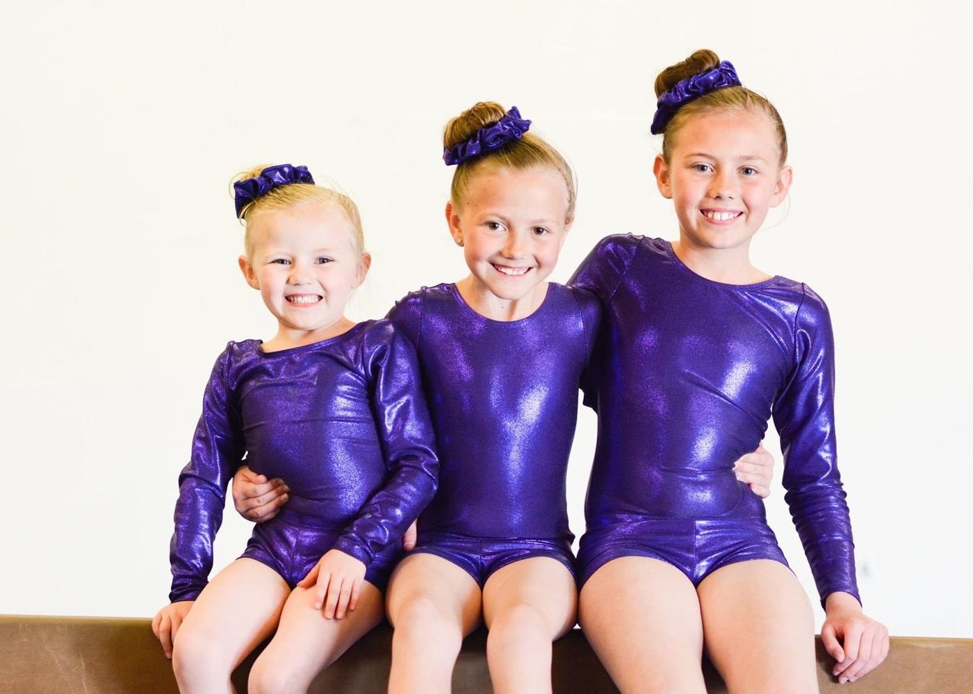 Three girls in matching purple leotards sitting together and smiling.
