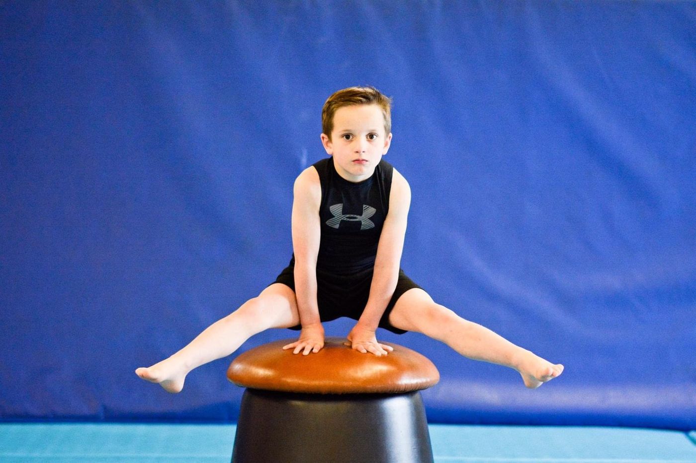 A young boy doing a straddle support on a mushroom apparatus against a blue backdrop.