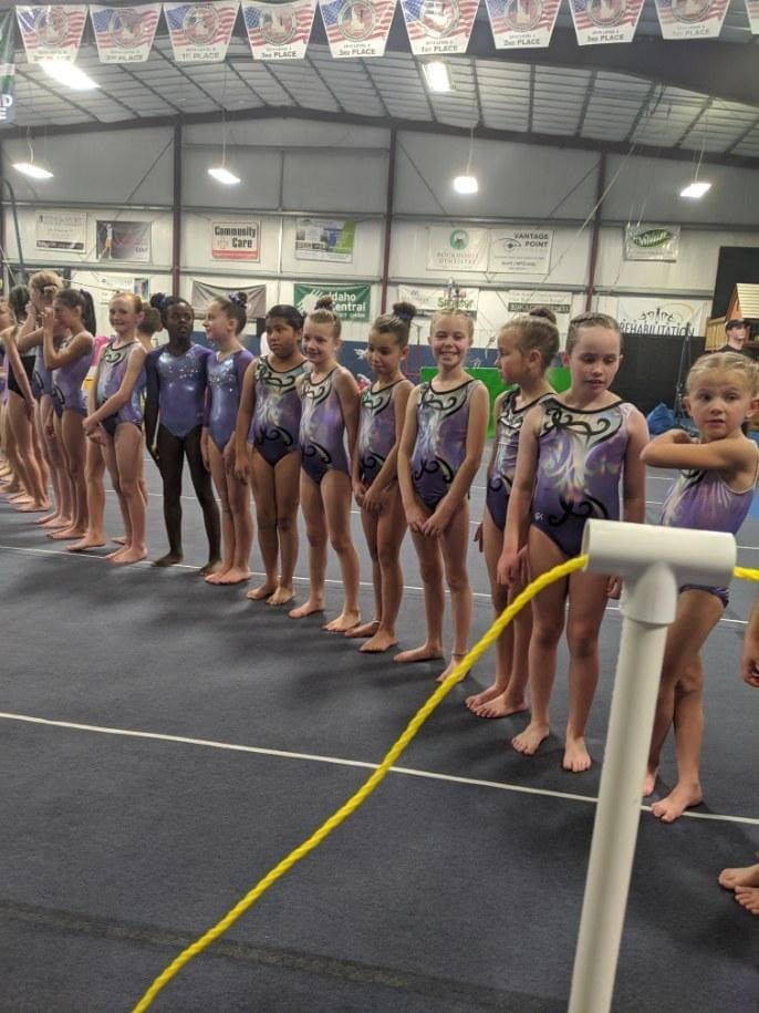 A long line of young gymnasts in purple leotards standing in the gym at a meet.