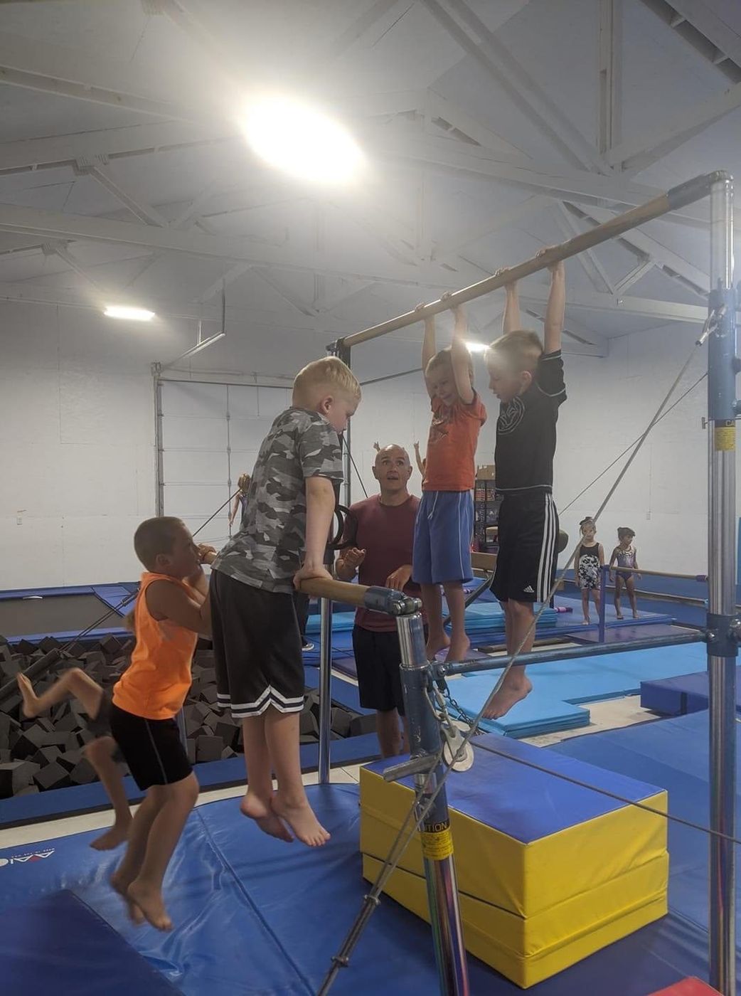 A male coach assisting boys swinging on the uneven bars in the gym.