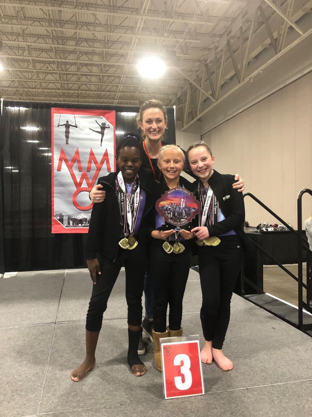 A coach posing with three gymnasts holding medals and a trophy at a competition.