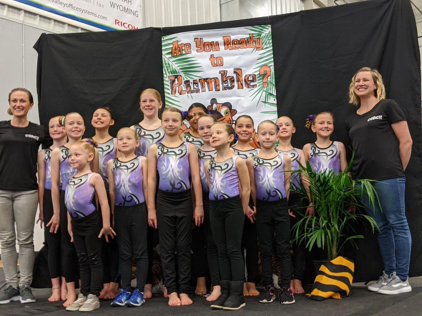 A team of young gymnasts and two coaches posing in front of an 'Are You Ready to Rumble' banner.