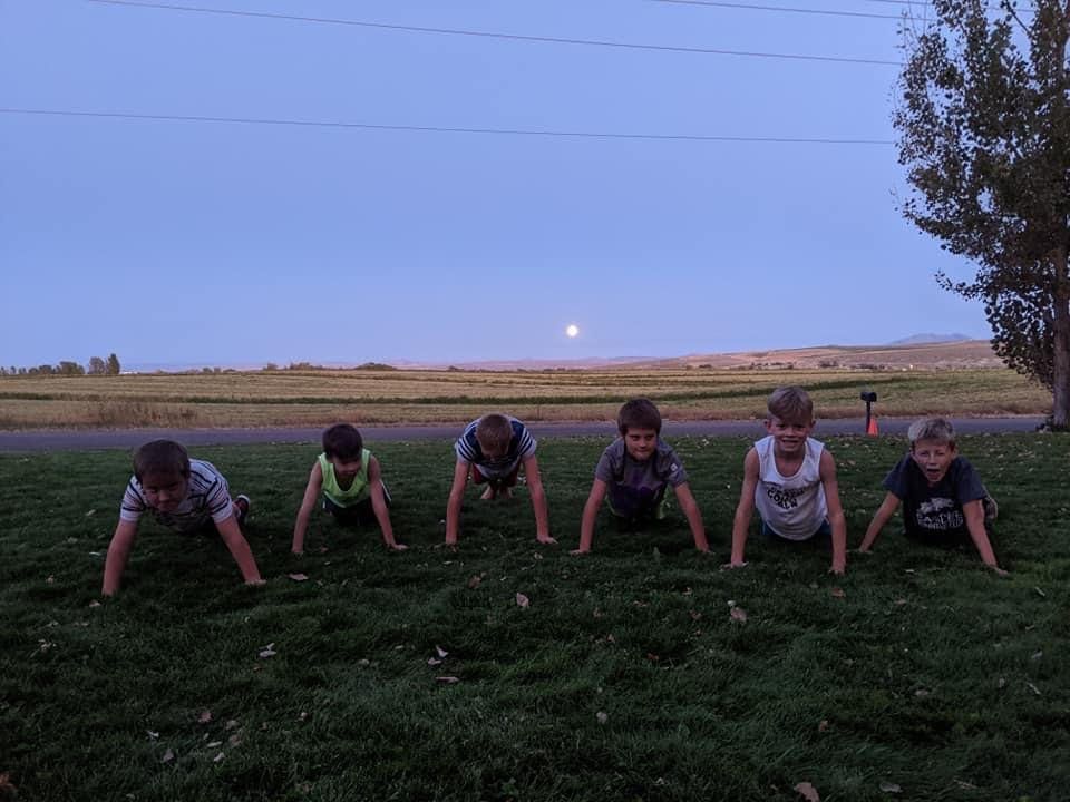 Six boys doing push-ups on a grassy field at dusk with a full moon behind them.