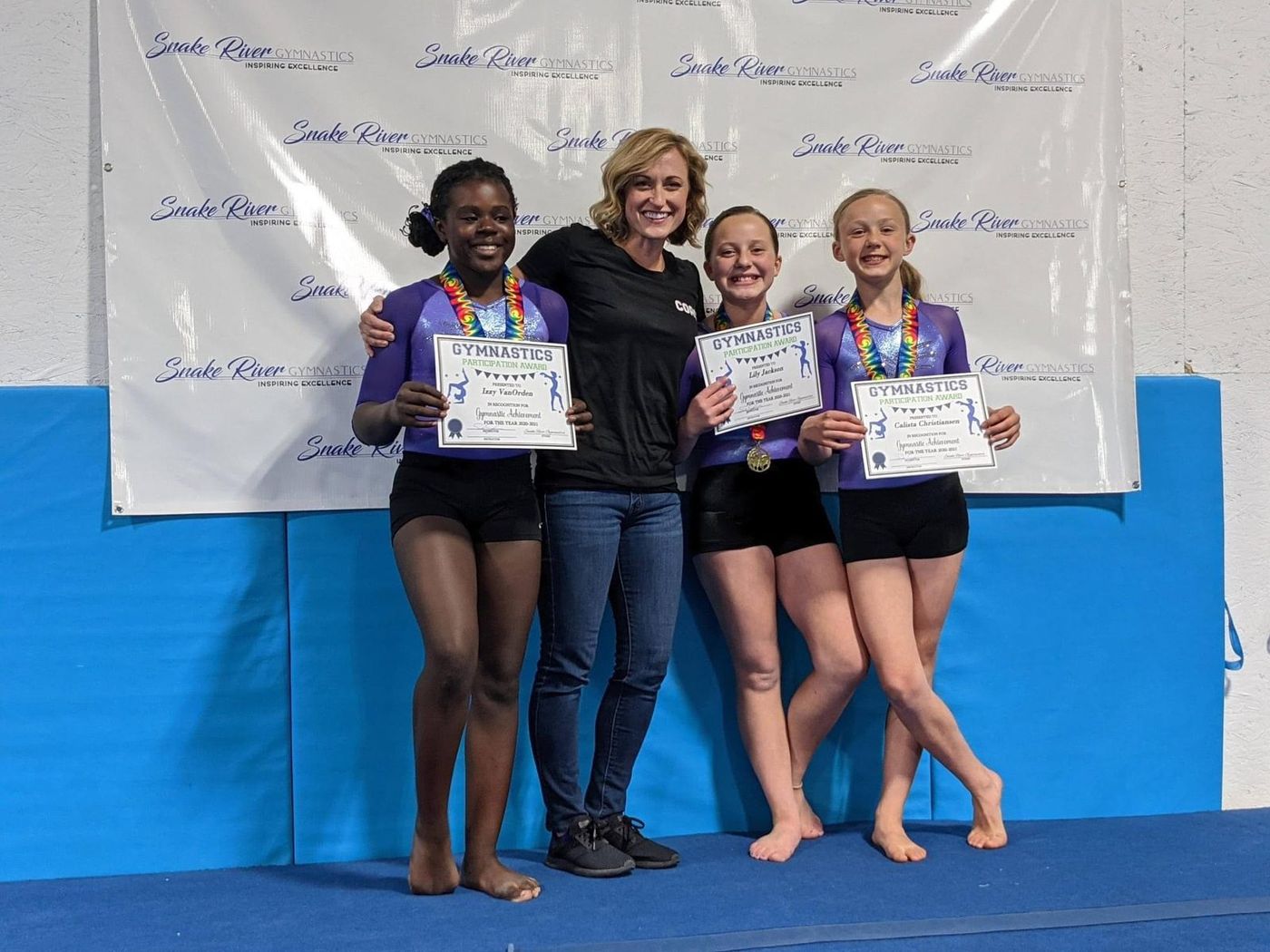 A coach posing with three gymnasts holding participation certificates in front of a Snake River Gymnastics banner.