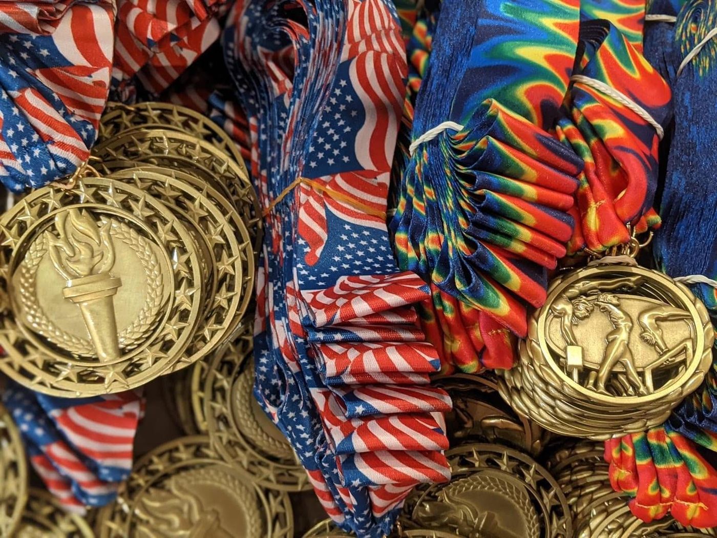 A close-up pile of gold medals with patriotic and tie-dye ribbons.