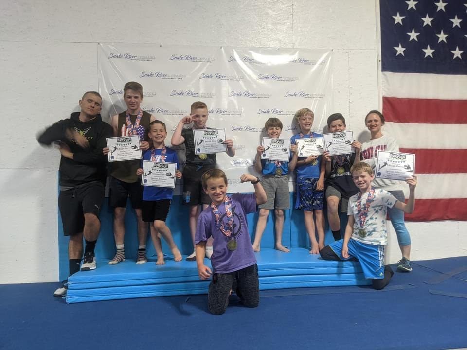 A group of boys holding certificates and medals with two coaches in front of a Snake River Gymnastics banner.