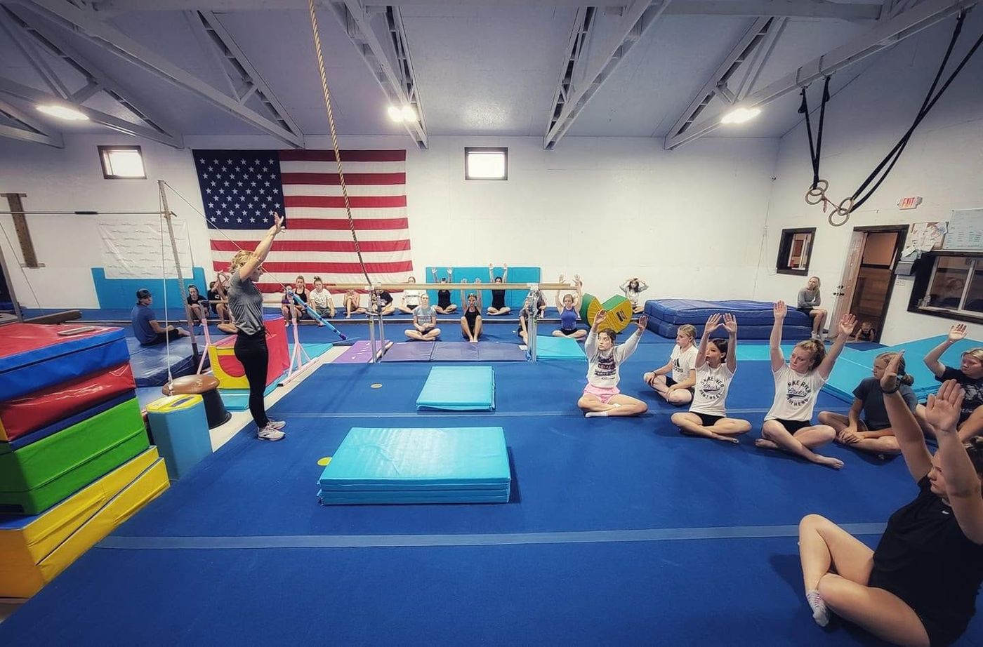 A coach leading a large group of gymnasts seated on the floor during a class in front of an American flag.