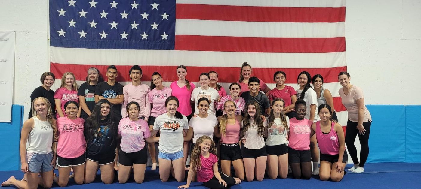 A large group of teen gymnasts and coaches in pink for a breast cancer awareness day in front of an American flag.