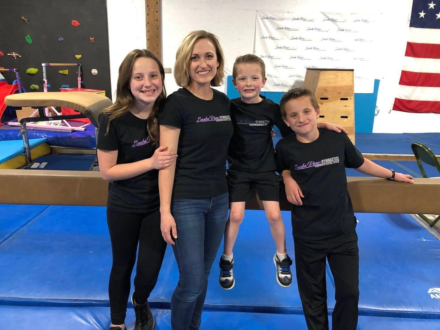 A coach posing with three children wearing Snake River Gymnastics shirts in the gym.