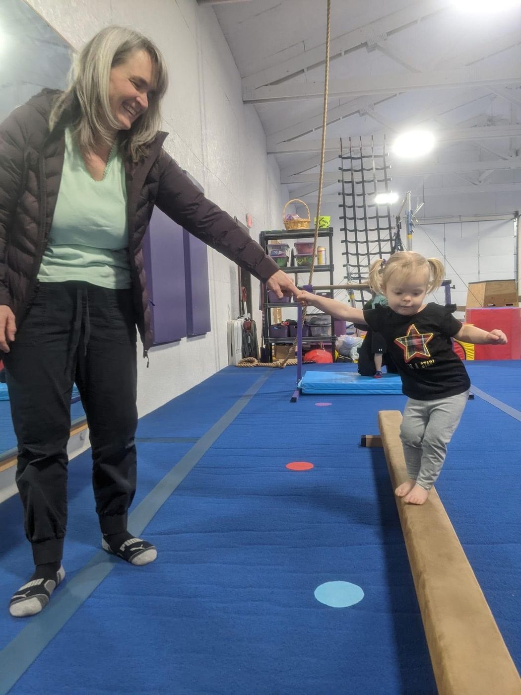 A coach helping a toddler balance on a low beam in the gym.