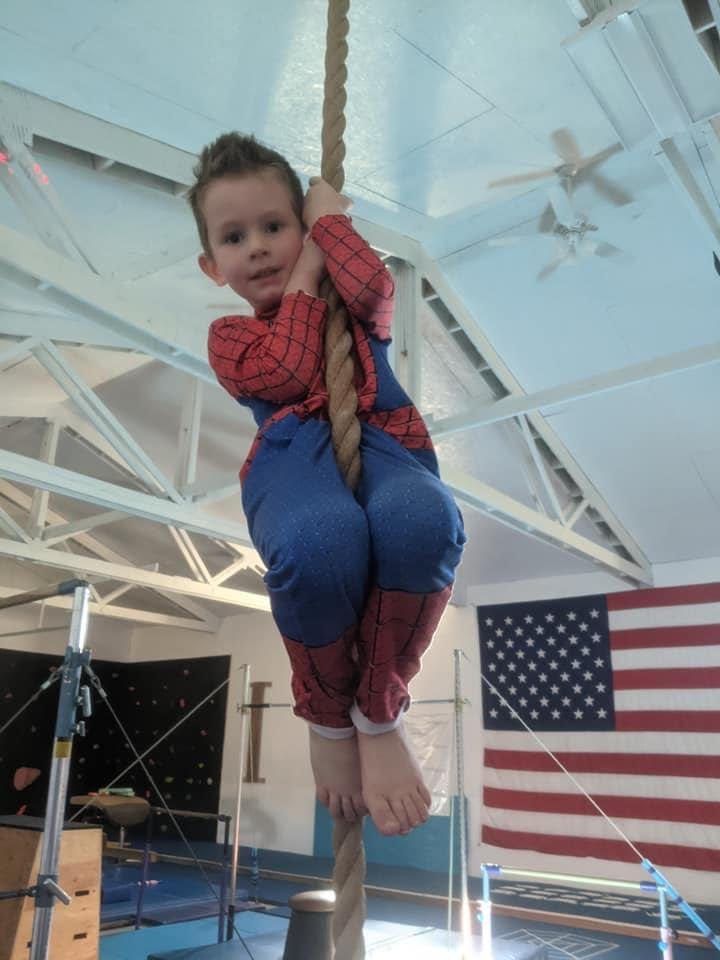 A young boy in a Spider-Man costume climbing a rope in the gym.