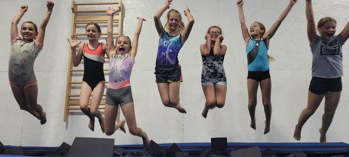 Seven girls jumping with arms raised into a foam pit in the gym.