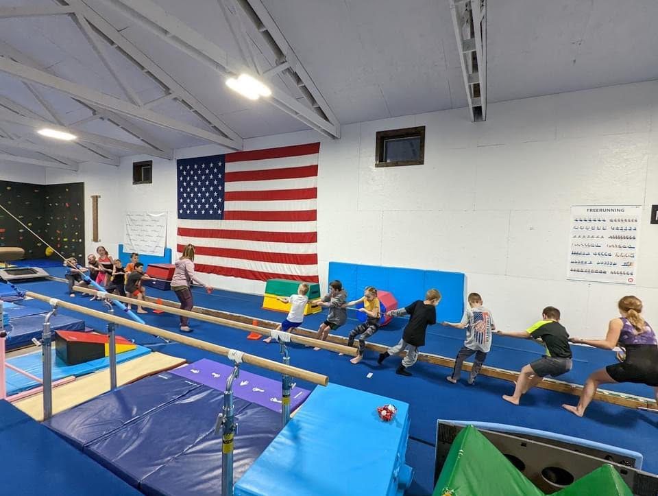 Children in a parkour class lined up doing a tug-of-war exercise on a bar in the gym with an American flag on the wall.