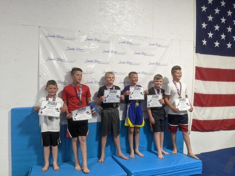 Six boys holding certificates and medals on the awards podium in front of a Snake River Gymnastics banner.