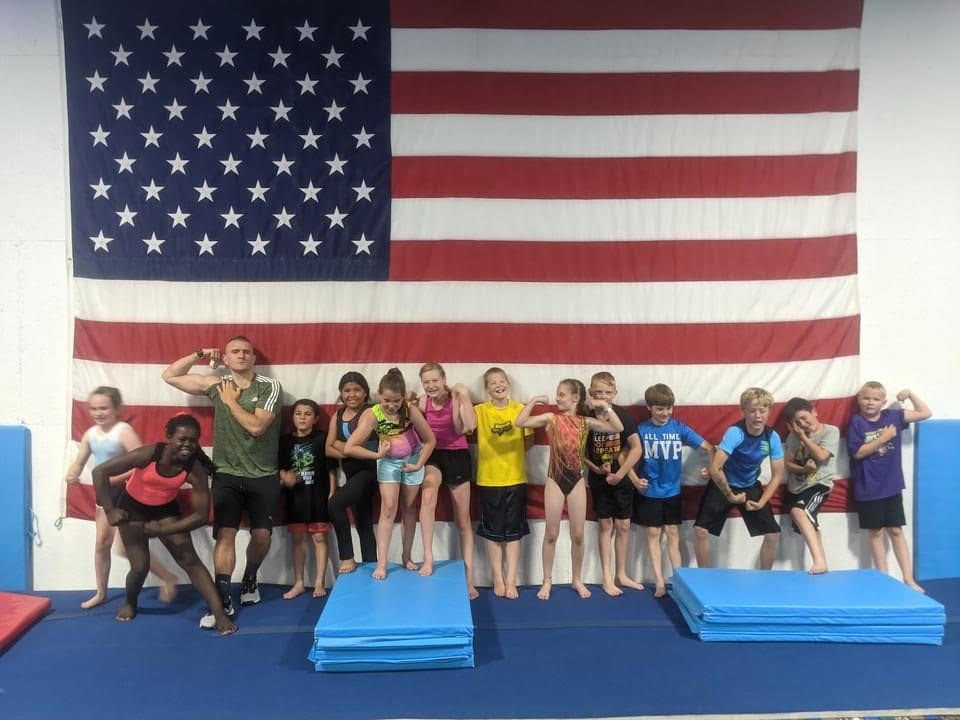 A group of kids and a coach flexing and posing in front of a large American flag in the gym.