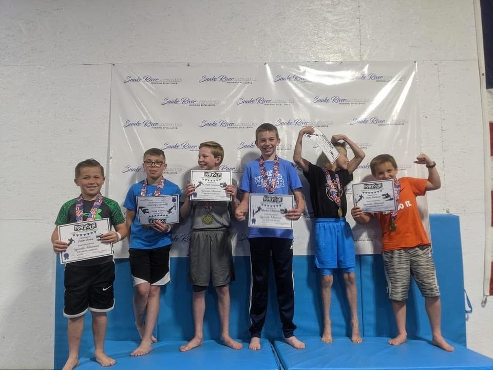 A group of boys holding certificates and medals posing in front of a Snake River Gymnastics banner.