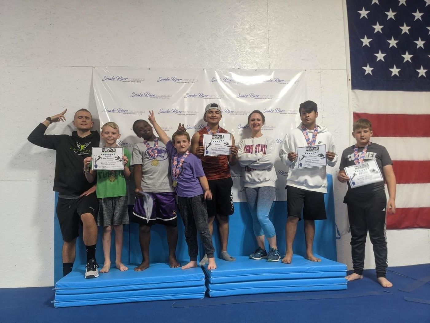 Boys and a coach holding certificates and medals on mats in front of a Snake River Gymnastics banner.