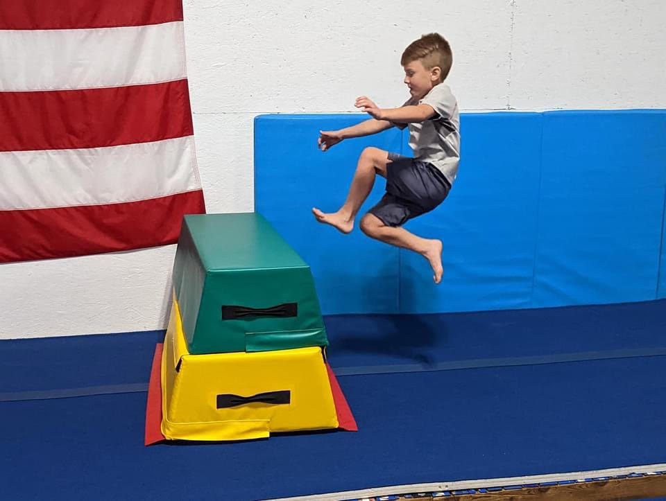 A young boy leaping off a colorful vaulting block in the gym with an American flag in the background.