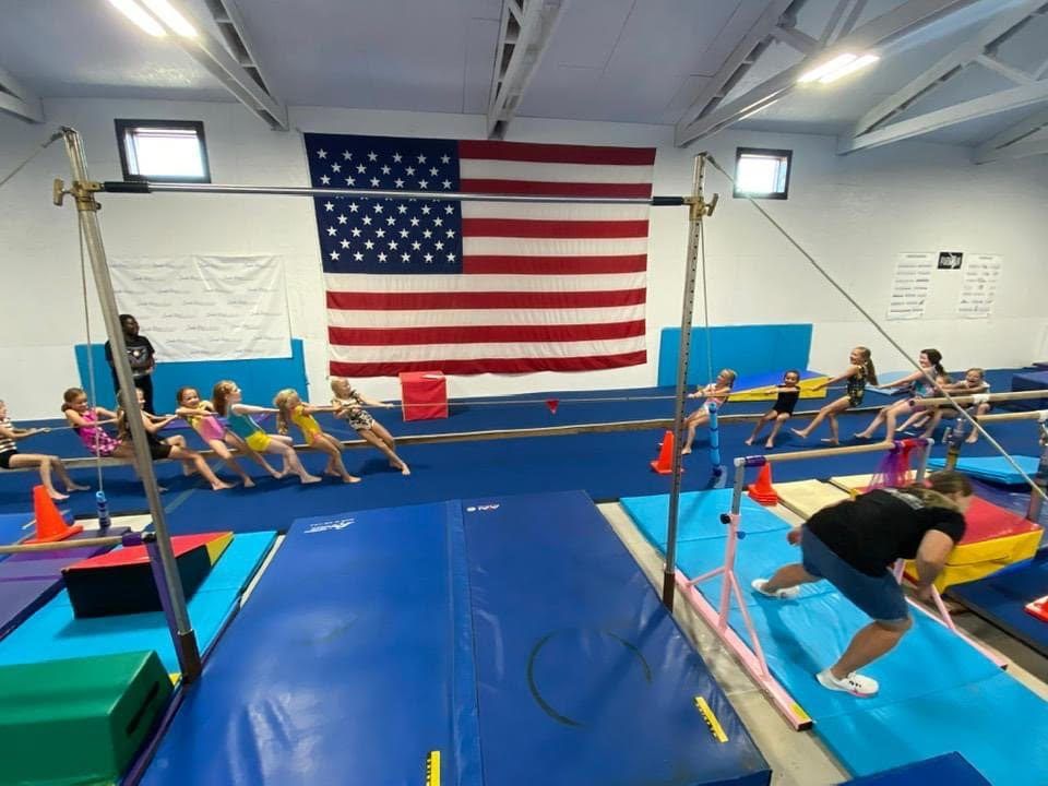 A group of young girls hanging from the bars during a class while a coach spots them near the American flag.