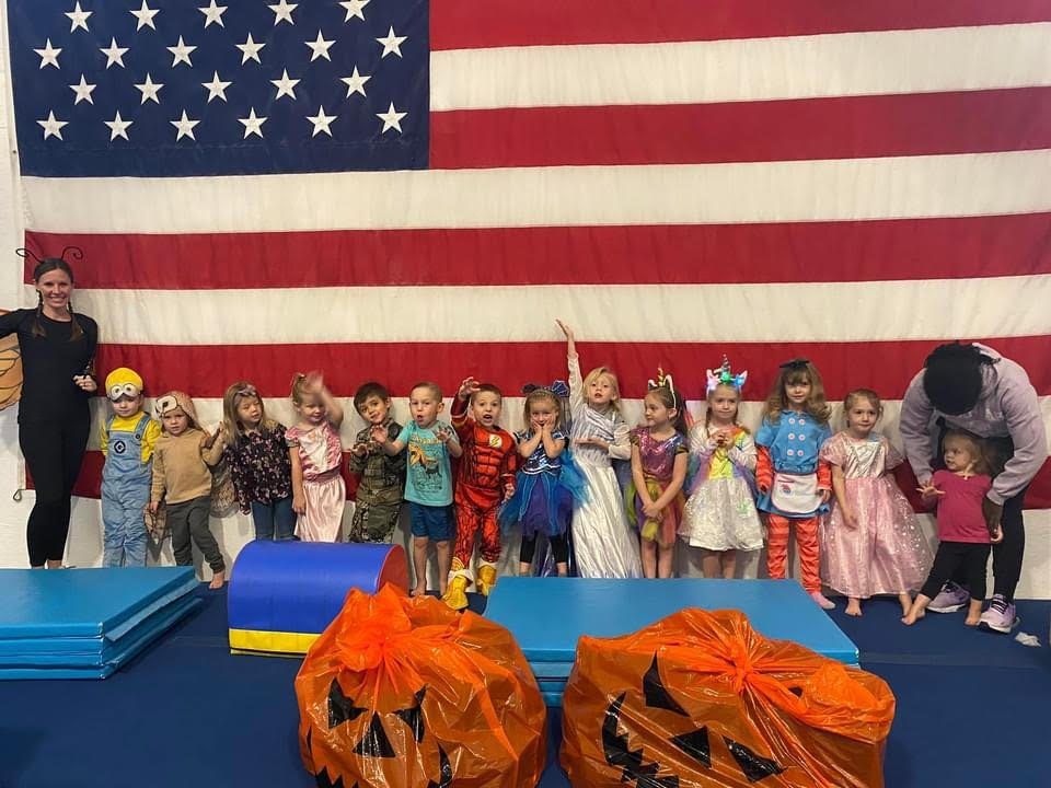 Young children in Halloween costumes lined up in front of an American flag with jack-o-lantern bags.