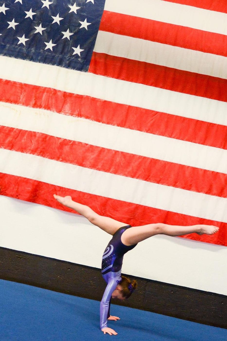 A gymnast holding a handstand on the floor in front of a large American flag.