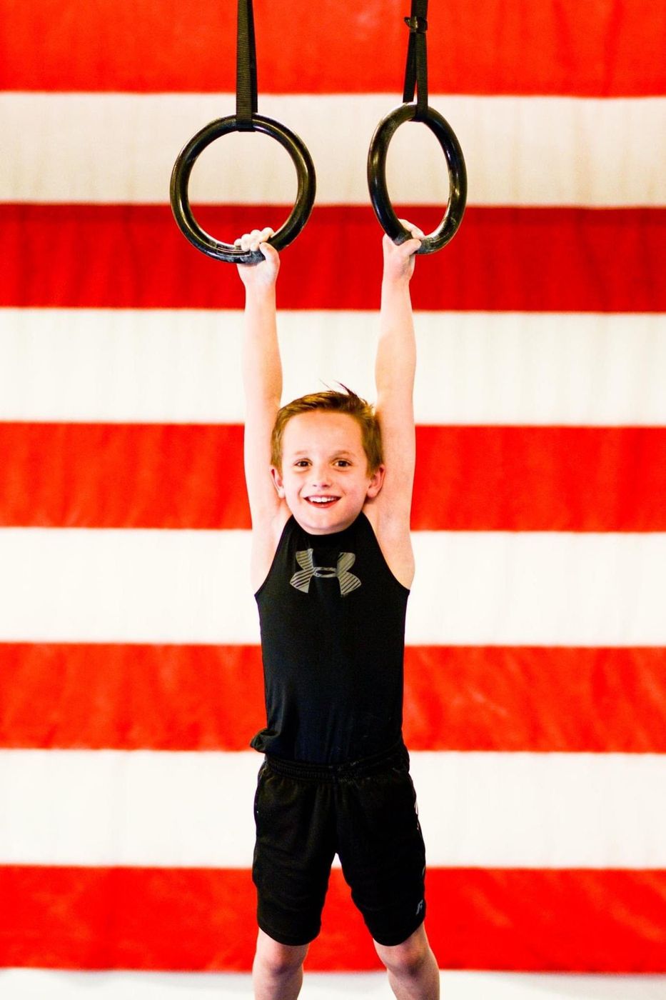 A smiling young boy hanging from the gymnastic rings in front of a striped flag backdrop.