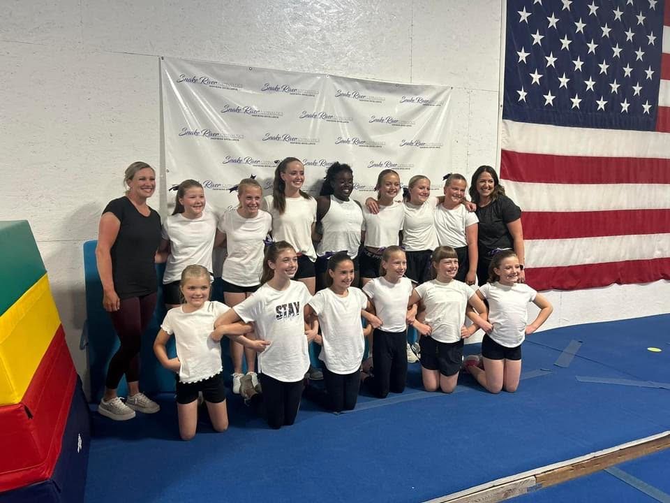 A cheer team in white shirts posing with two coaches in front of a Snake River Gymnastics banner and American flag.