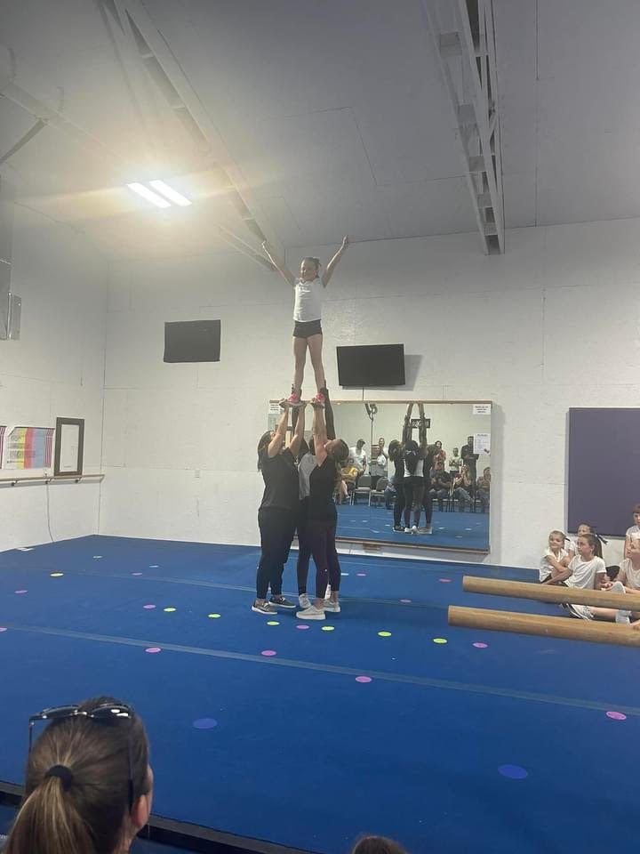 Spotters supporting a young cheerleader in a standing stunt during a gym showcase.