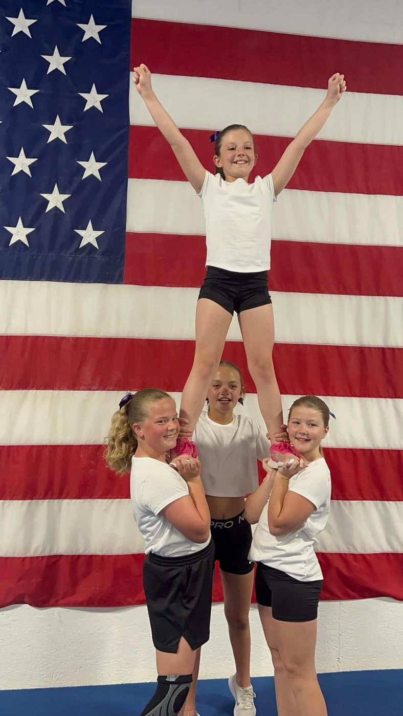 Three young cheerleaders holding up a teammate in a stunt in front of an American flag.