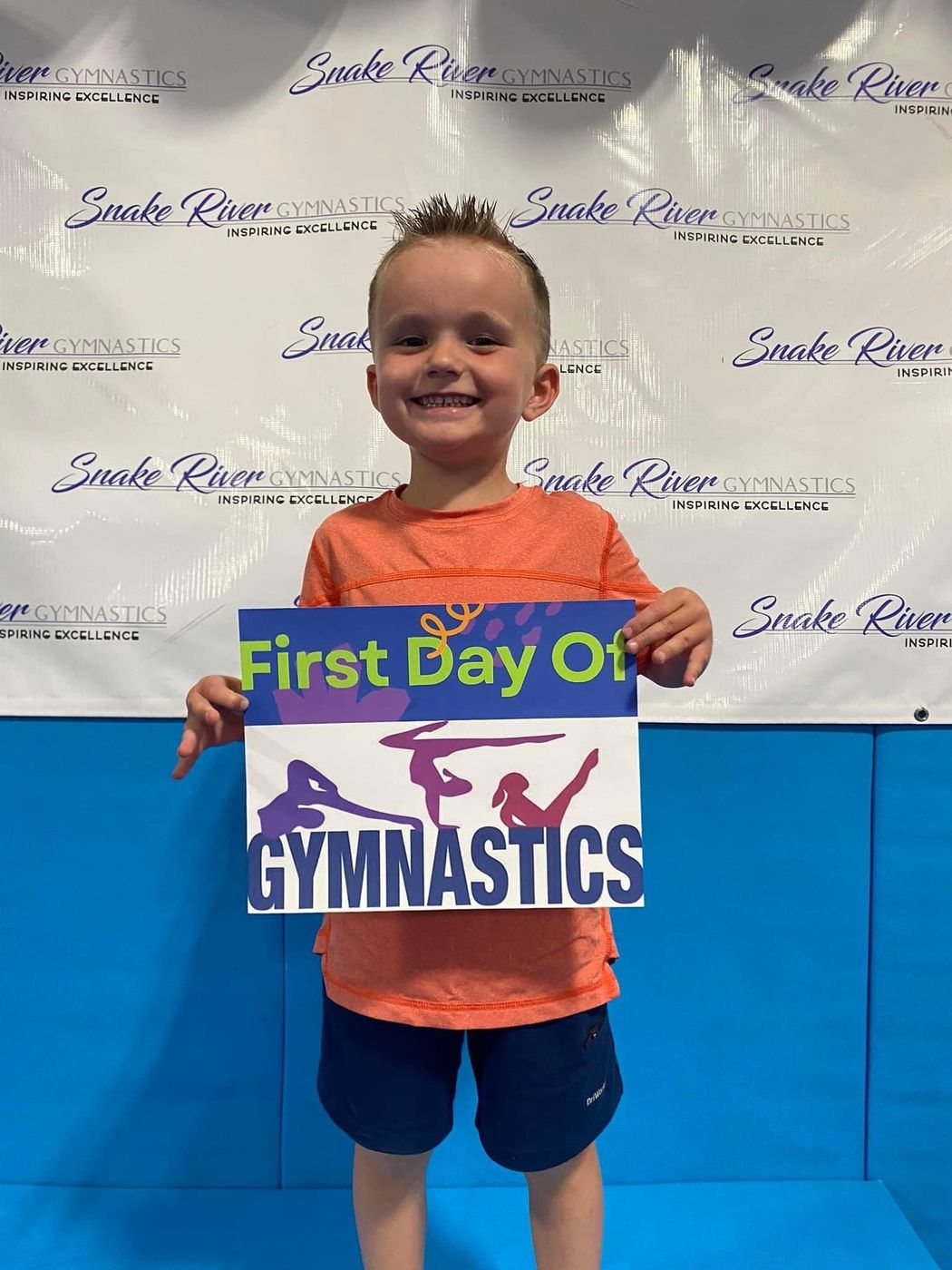 A smiling boy holding a First Day of Gymnastics sign in front of a Snake River Gymnastics backdrop.