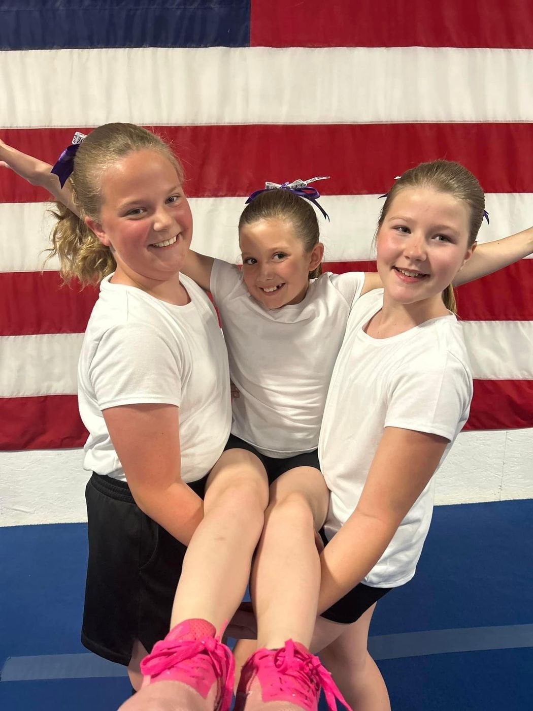 Two girls carrying a younger teammate while smiling in front of an American flag.