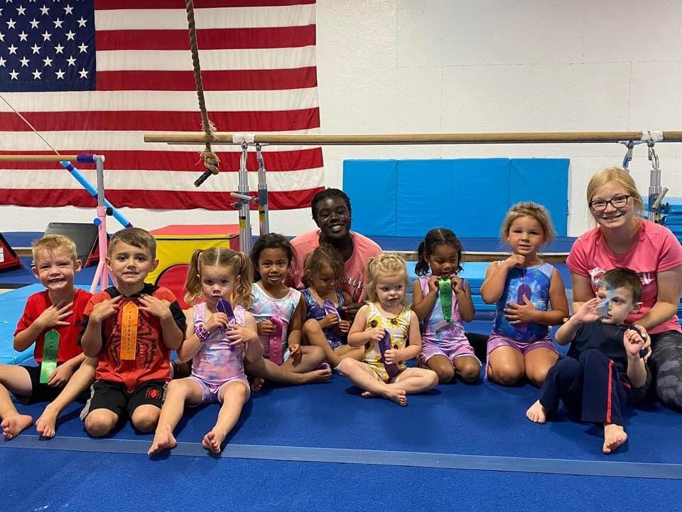 A group of young children holding ribbons with two teen coaches in front of an American flag.