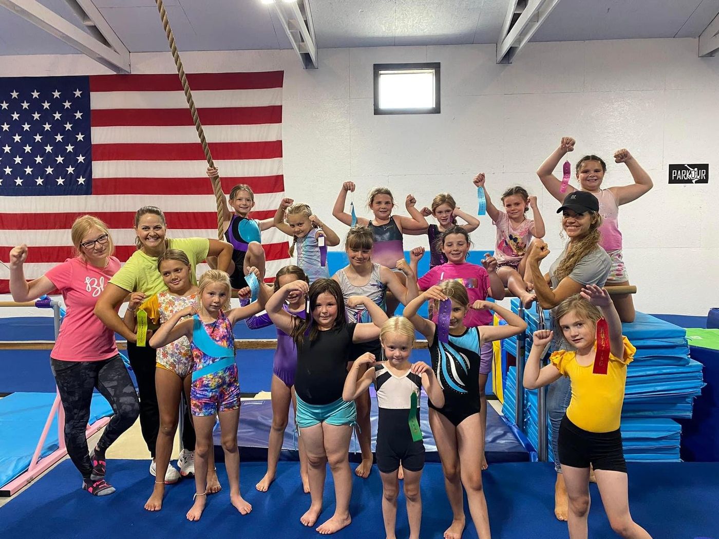 A large group of girls and coaches flexing and posing together in the gym near an American flag.
