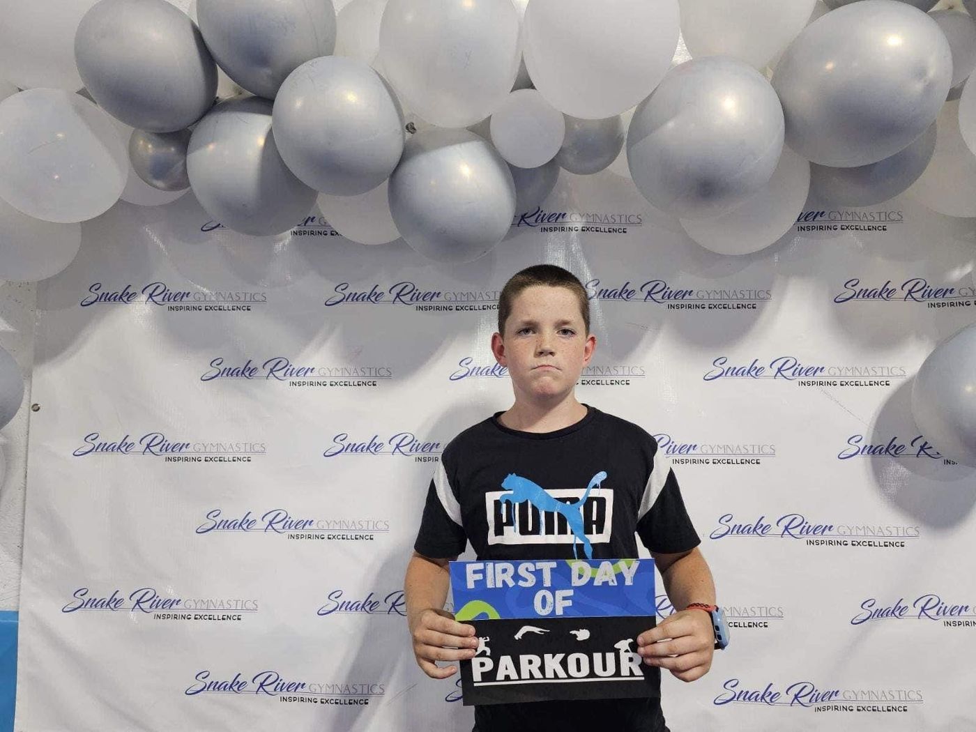 A boy holding a First Day of Parkour sign in front of a Snake River Gymnastics balloon backdrop.