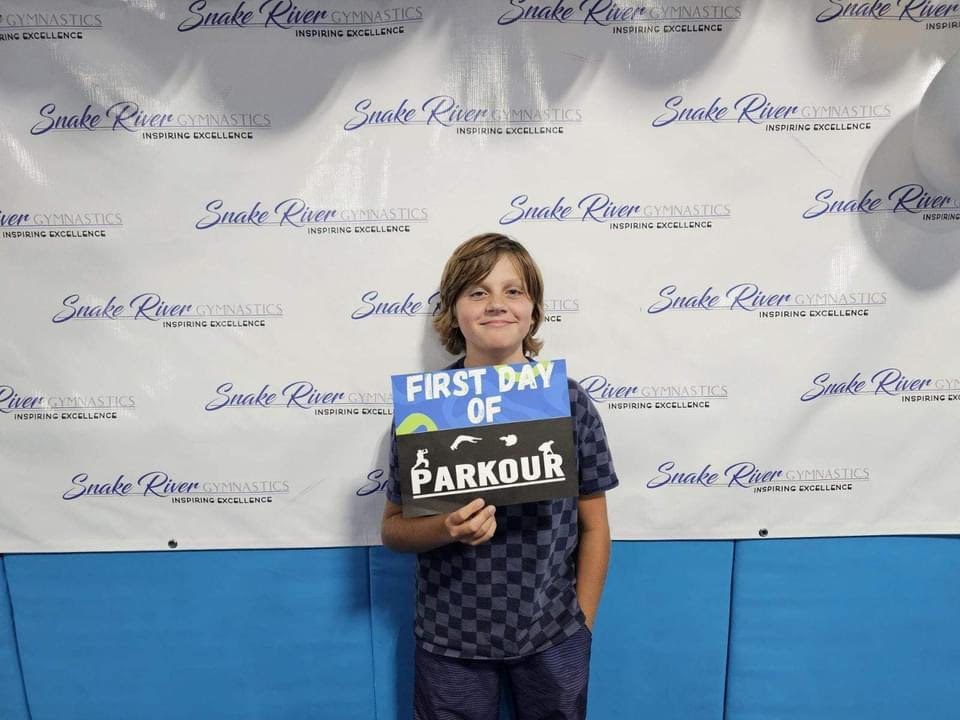 A boy holding a First Day of Parkour sign in front of a Snake River Gymnastics backdrop.