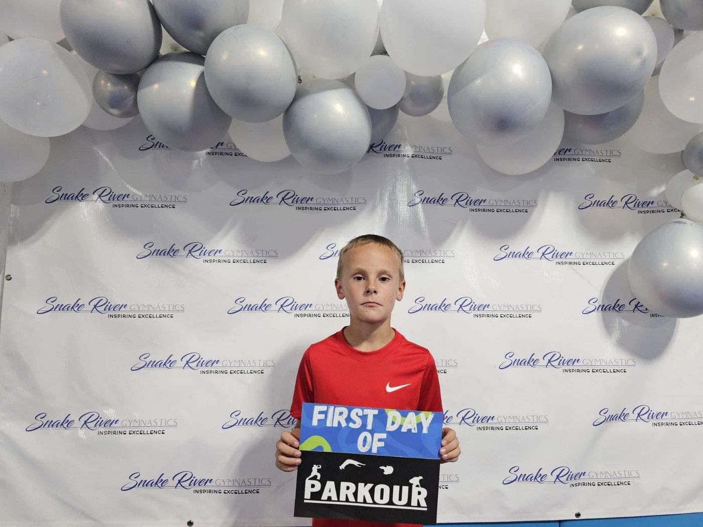 A boy in a red shirt holding a First Day of Parkour sign in front of a balloon backdrop.