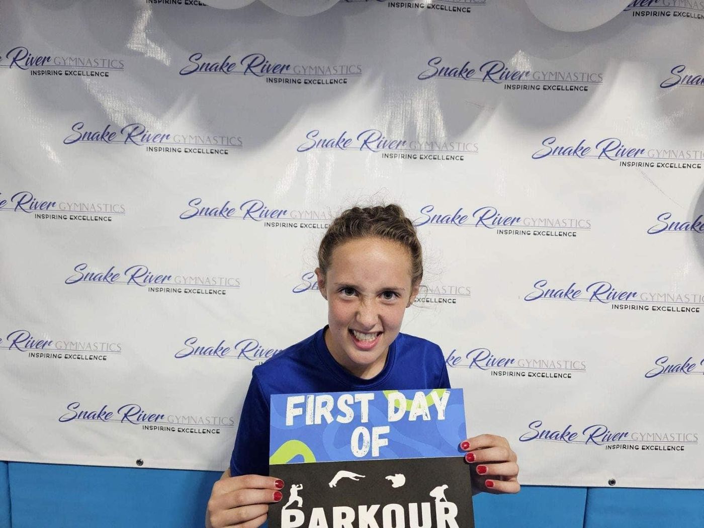 A girl holding a First Day of Parkour sign in front of a Snake River Gymnastics backdrop.
